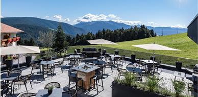 Sonnige Panoramaterrasse mit Sitzplätzen, Sonnenschirmen und Blick auf grüne Wiesen und Berge.