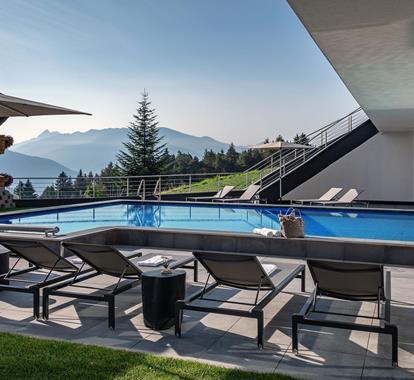 Moderner Außenpool mit stilvollen Liegen, Sonnenschirmen und Panoramablick auf bewaldete Berge und Täler in der Morgensonne.