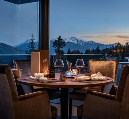 Stimmungsvoll gedeckter Tisch mit Wein und Kerzenlicht vor Panoramablick auf die Berge bei Sonnenuntergang.