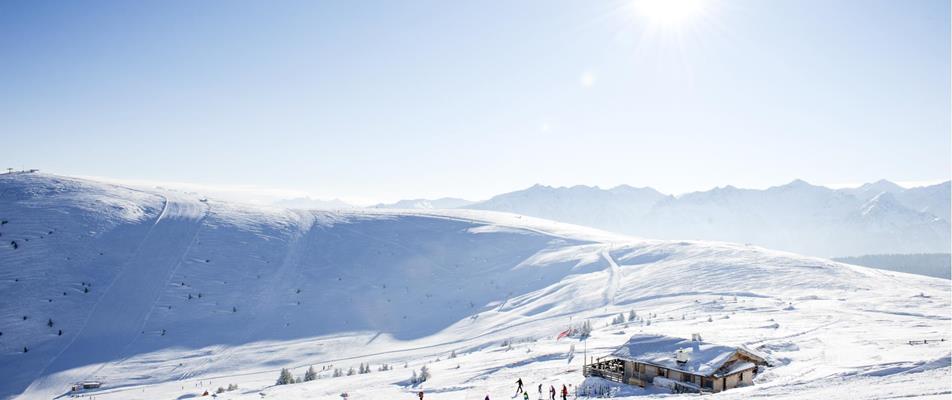 Snowy winter landscape with a cabin, skiers, and bright sunshine in the Dolomites.