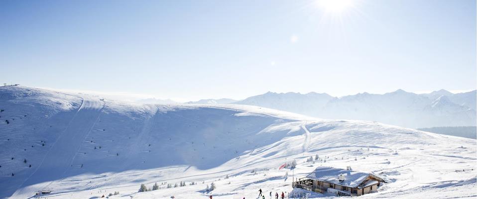 Verschneite Winterlandschaft mit Hütte, Skifahrern und Sonnenschein in den Dolomiten.