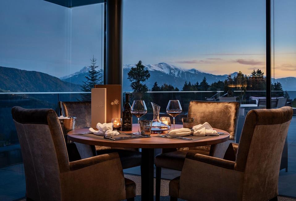 Stimmungsvoll gedeckter Tisch mit Wein und Kerzenlicht vor Panoramablick auf die Berge bei Sonnenuntergang.