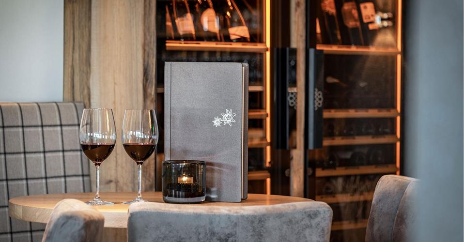 Two glasses of red wine, a menu, and a lit candle on a round wooden table in front of an illuminated wine cabinet.