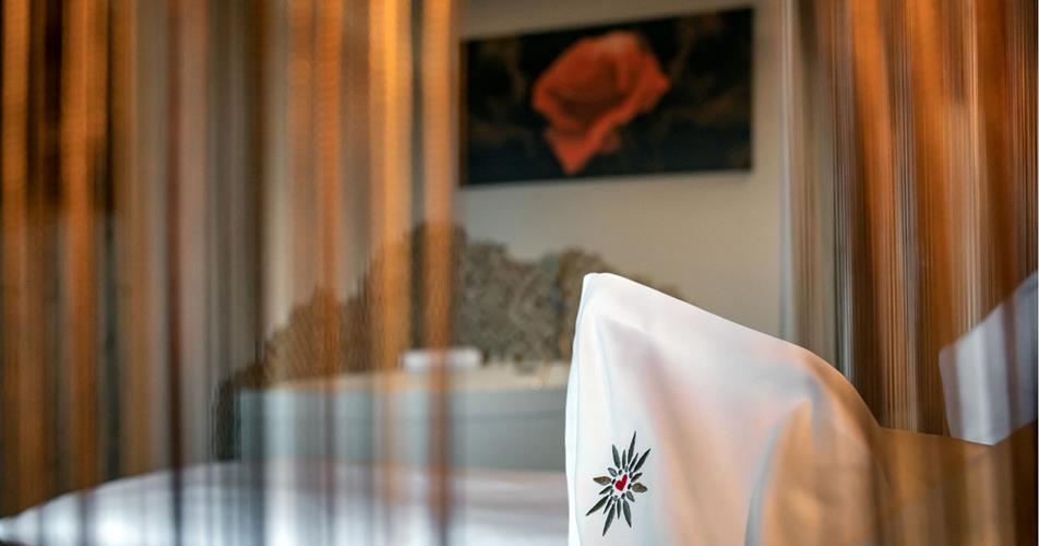 View through a semi-transparent curtain of a bed with an edelweiss-embroidered pillow; a painting of a red rose hangs in the background.