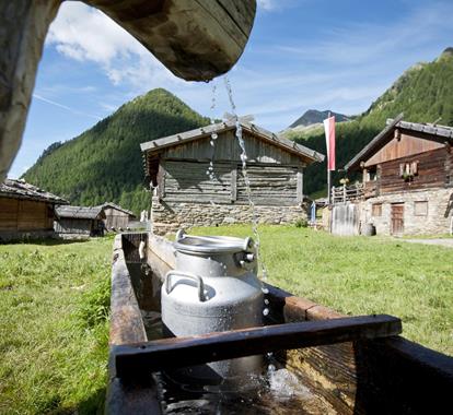Water trough with milk churns in a mountain village, surrounded by traditional wooden huts and peaks.
