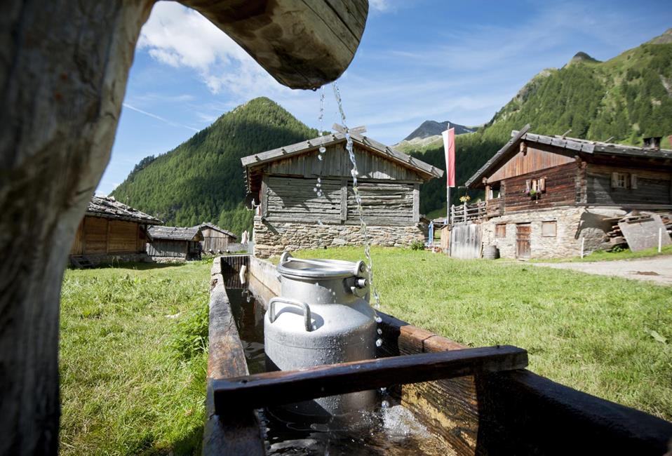 Wassertränke mit Milchkannen in einem Bergdorf, umgeben von traditionellen Holzhütten und Bergen.