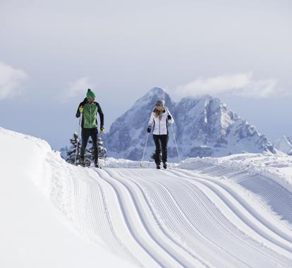 Due sciatori di fondo su pista appena battuta con maestose montagne sullo sfondo.
