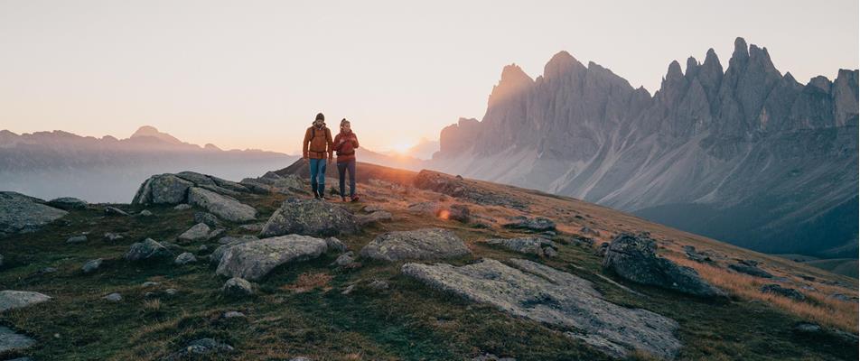 Two hikers in warm clothing walk at sunset across a rocky mountain slope with jagged Dolomite peaks in the background.