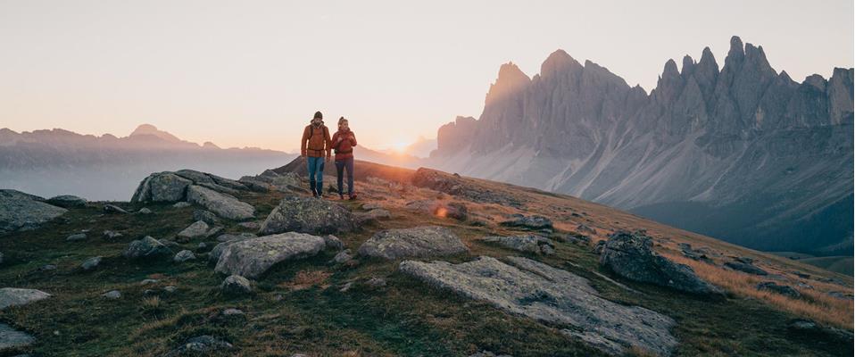 Two hikers in warm clothing walk at sunset across a rocky mountain slope with jagged Dolomite peaks in the background.