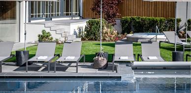 Zona esterna elegante con lettini moderni, ombrelloni e giardino curato accanto alla piscina; sullo sfondo una vasca idromassaggio e architettura raffinata.