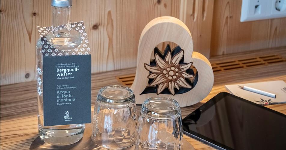 Karaffe mit Südtiroler Bergquellwasser und zwei Gläsern auf Holztablett, daneben ein Herz aus Holz mit Edelweißmotiv.