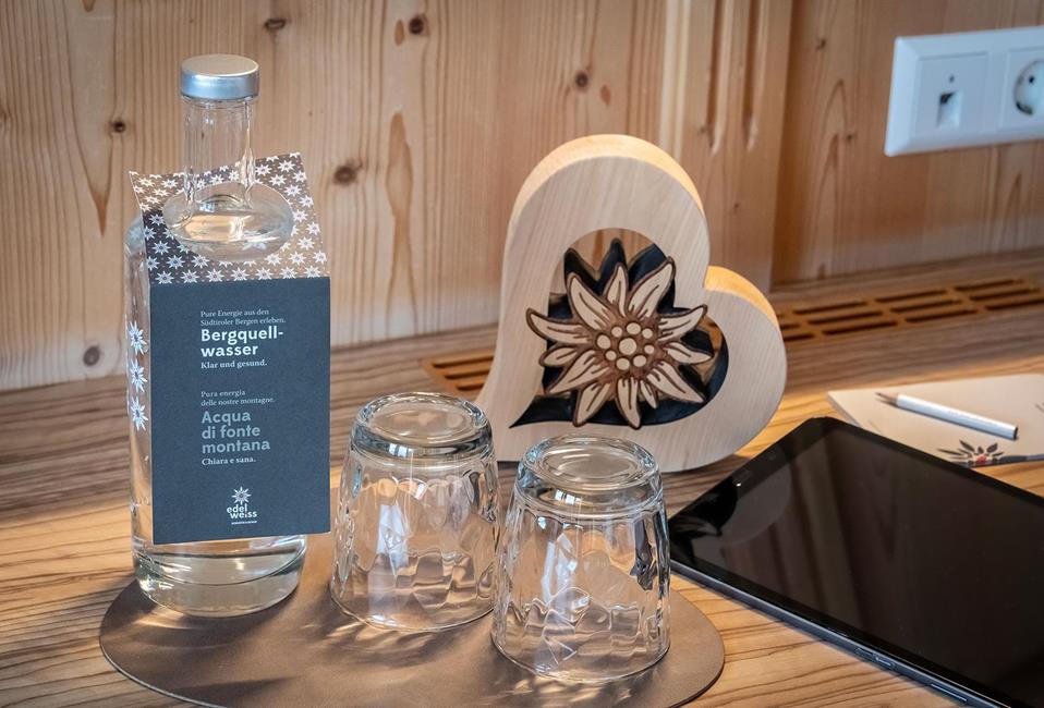 Caraffa con acqua di sorgente alpina dell'Alto Adige e due bicchieri su vassoio in legno, accanto un cuore in legno con stella alpina.