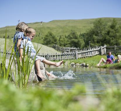 Kids play in a creek with their feet in the water, people relax on the grass in the background.