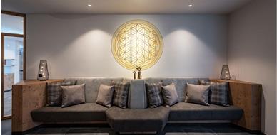 Modern relaxation area with cushioned bench seating, decorative pillows, and soft lighting; a glowing mandala featuring the Flower of Life on the wall.