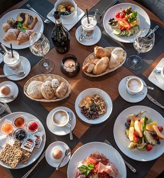 Ricca colazione con pane, salumi, frutta fresca, cereali, caffè e succhi su tavola apparecchiata.