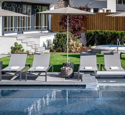 Elegant outdoor area with modern sun loungers, umbrellas, and a well-kept garden by the poolside; a hot tub and stylish architecture in the background.