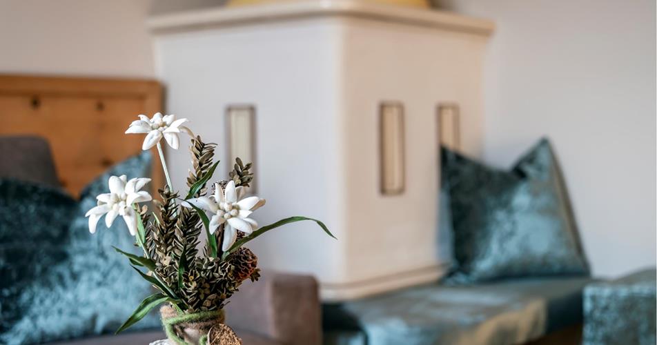 Close-up of an edelweiss arrangement in a rustic vase with a heart ornament; in the background a tiled stove and velvet cushions.