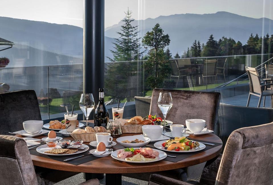 Reichhaltiges Frühstück mit frischen Brötchen, Obst, Aufschnitt und Heißgetränken mit Bergblick.
